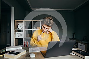Young male student studying at home with books and laptop on bedroom background, looking at notebook with serous face and biting