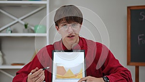 A young male student wearing glasses gives a presentation of his work, sitting at a table indoors at home. The guy