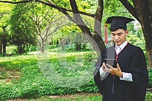 Young male students dressed in black graduation gown. Nature as a background. Boy cheerfully smiling, looking at mobile phone