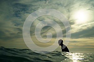 A young male surfer in silhouette sits waiting for a wave at Piha Beach, New Zealand