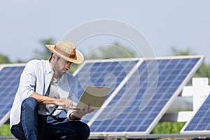 young male technician in sun hat and shirt holding laptop and checking solar panels while standing near farm