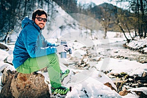 Young male tourist drinks hot coffee from a thermos near a river in the mountains.