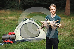 A young male tourist holding woods for a fire near tent in the forest