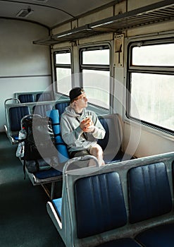 Young male tourist sits on a train while traveling on the railway with a backpack, uses a smartphone and looks out the window