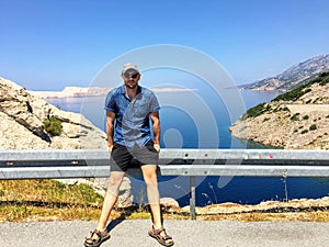 A young male tourist sitting along the railing at a highway stop along the adriatic highway in Croatia.  It is a beautiful sunny w