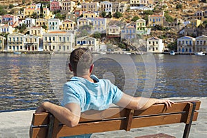 Young male tourist sitting on a bench and looking at the port of Symi, with many different color