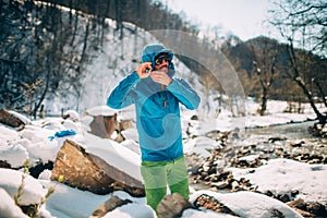 Young male tourist straightens his jacket near a river in the mountains.