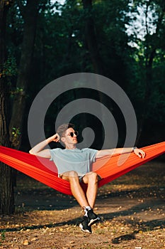 Young male tourist in sunglasses and light clothes resting sitting on a hammock in the woods at sunset, looking away. Vertical