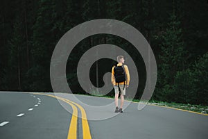 Young male traveler walking down the asphalt road in the evening looking at the mountain coniferous forest. Tourist guy in yellow