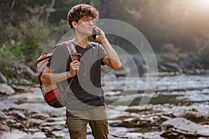 Young male traveler walking in mountains at river and talking on mobile phone. Hiking.