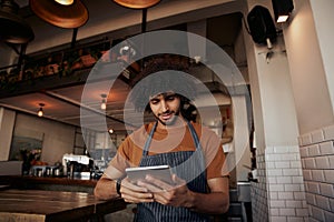 Young male waiter wearing apron uniform in cafe using digital tablet to watch recipe