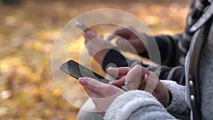 Young male and woman use phone in the autumn park sitting on a bench. Focus on the girl hands