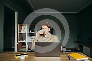 Young male worker in shirt working at home remotely on laptop and drinking hot beverage from cup. Freelancer in wireless