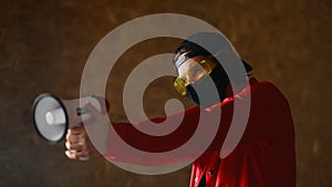 A young man of 25-30 years old in a black protective mask, yellow goggles, a cap and a red jacket holds a megaphone