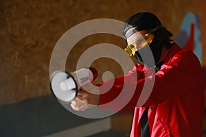 A young man of 25-30 years old in a black protective mask, yellow goggles, a cap and a red jacket holds a megaphone