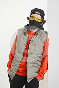 A young man of 25-30 years old in a black protective mask, yellow goggles, a cap and a red jacket posing on a gray background.