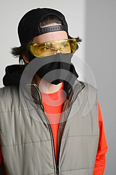 A young man of 25-30 years old in a black protective mask, yellow goggles, a cap and a red jacket posing on a gray background.