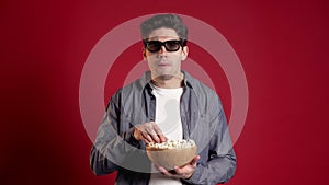 Young man in 3d glasses watching fascinating comedy or horror movie and eating popcorn on red studio background.