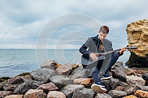 Young man with acoustic guitar playing on beach surrounded with rocks on rainy day