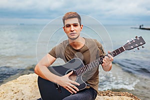 Young man with acoustic guitar playing on beach surrounded with rocks on rainy day