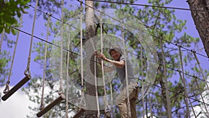 A young man in an adventure park. He wears a safety harness. He climbs on a high rope trail. Outdoor amusement center