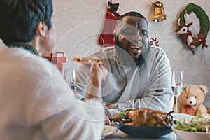 Young man with african american husband enjoying christmas lunch of turkey at decorated home with stockings, candy cane