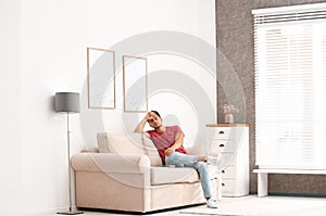 Young man with air conditioner remote control suffering from heat on sofa