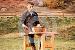 Young man in apron sit at potters wheel, moulding wet clay on spinning turntable outdoor in summer