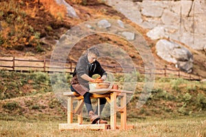 Young man in apron sit at potters wheel, moulding wet clay on spinning turntable outdoor in summer