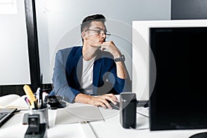 Young man architect in glasses dressed in a business suit sits at a desk in front of a computer in the office