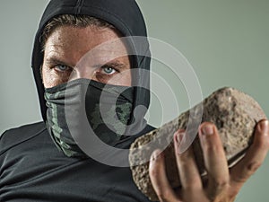 Young man as radical and aggressive anarchist rioter holding brick . furious anti-system protester in face mask throwing stone in