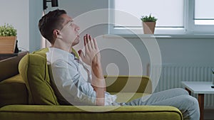 Young man asking for help about problem, praying to God. Religious male sit on sofa at home put hands in prayer. Guy