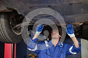 Young man auto mechanic repairman wearing eyeglasses in uniform checking car suspension repair in auto garage, service and