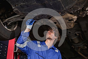 young man auto mechanic repairman wearing eyeglasses in uniform checking car suspension repair in auto garage, service and