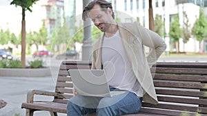 Young Man with Back Pain Using Laptop while Sitting on Bench