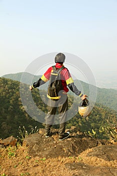 Young man with backpack and helmet standing with raised hands on top of a mountain and enjoying valley view