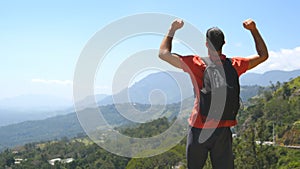 Young man with backpack reaching up top of mountain and raised hands. Male tourist standing on the edge of beautiful