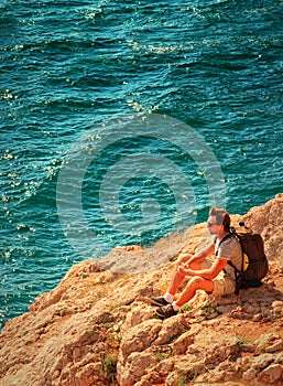 Young Man with backpack relaxing on rocky cliff with blue Sea on background