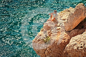 Young Man with backpack relaxing on rocky cliff with blue Sea on background