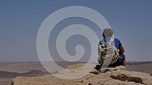 Young man with backpack sitting on cliff`s edge and looking at the desert
