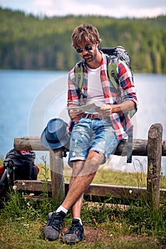 Young man with backpack sitting on fence in nature in front of lake looking at map. Sumeer hiking. Recration, lifestyle, nature