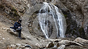 young man with backpack sitting near the mountain river waterfall