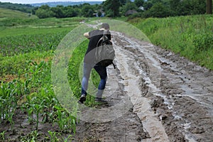 A young man with a backpack is trying to walk on a wet dirt road in heavy rain. Travel difficulties