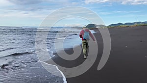 Young man with a backpack walks along the black sand beach and runs away from the rolling waves.