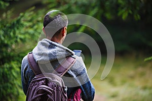 A young man with a backpack in the woods looking at the phone-a view from the back.