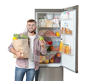 Young man with bag of groceries and apple near open refrigerator