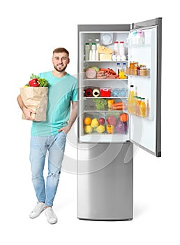 Young man with bag of groceries near open refrigerator
