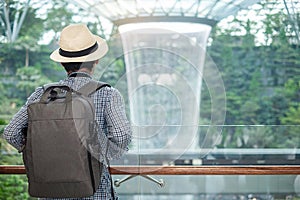 Young man with bag and hat, Asian traveler standing and looking to beautiful rain vortex at Jewel Changi Airport, landmark and