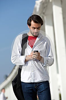 Young man with bag walking and sending text message outdoors