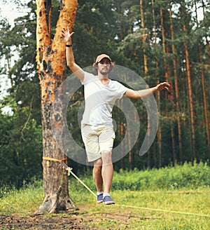 young man balancing his arms walk on a loose rope tied between two trees, male training slack rope walking, slacklining outdoors